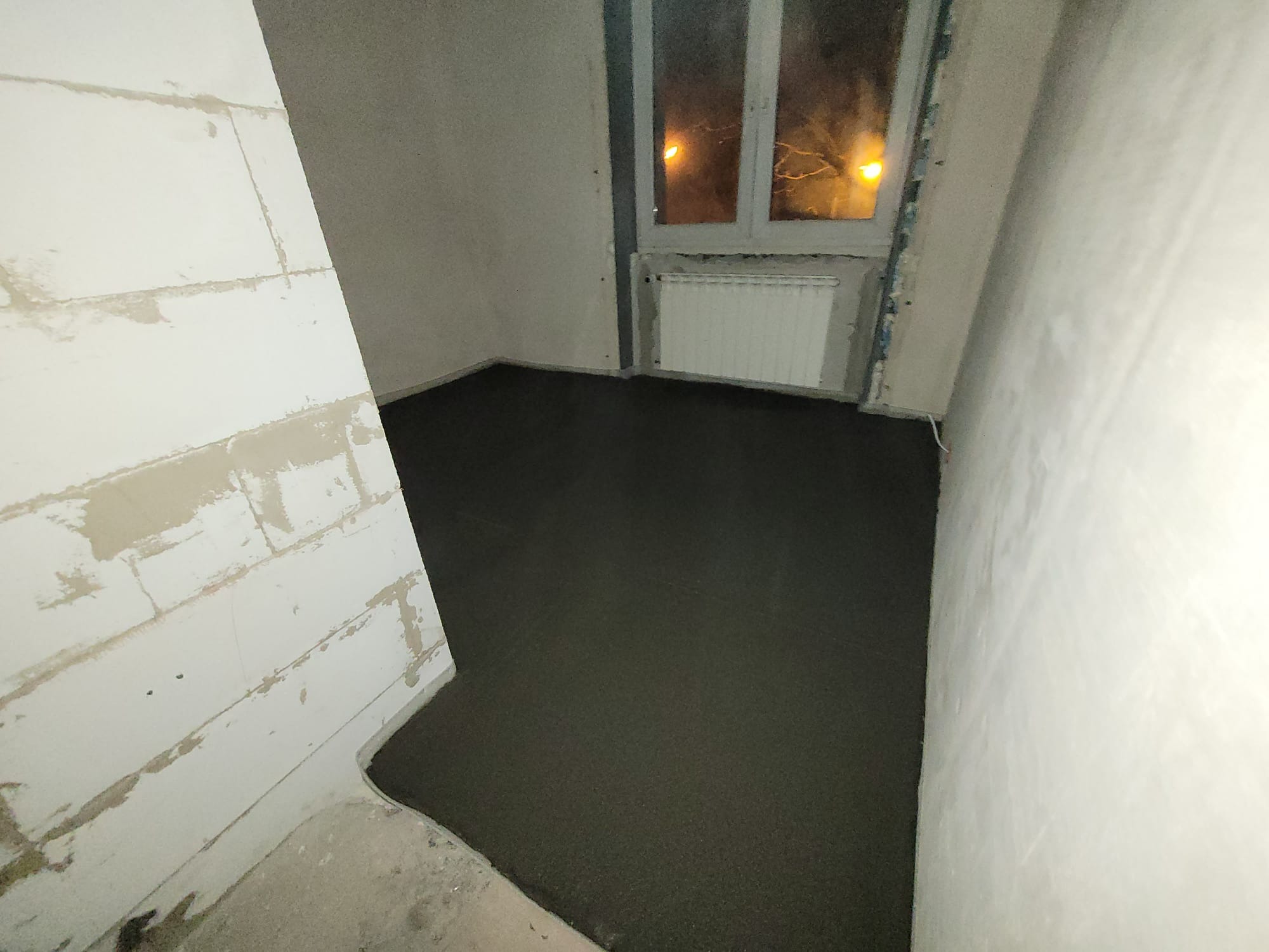 Wylana, ciemna posadzka w surowym wnętrzu. Widoczny grzejnik pod oknem i ściany w stanie deweloperskim. Perspektywa z narożnika pomieszczenia.