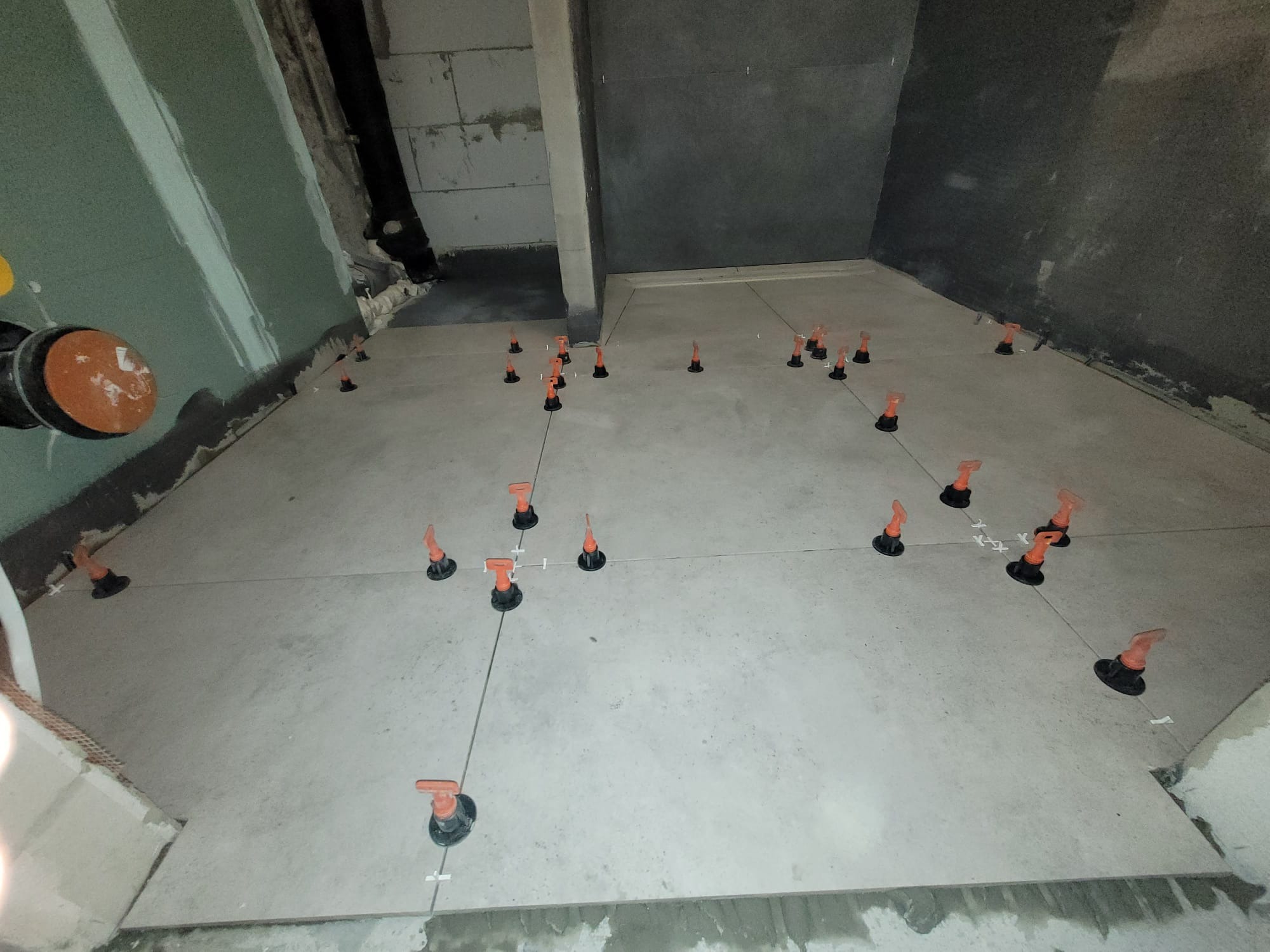 Układanie płytek podłogowych z użyciem systemu poziomowania. Widoczne pomarańczowe kliny i czarne podstawki na jasnych płytkach w surowym wnętrzu.