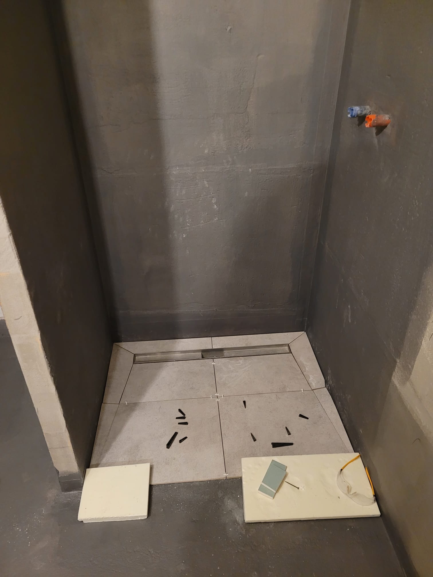 Wykładanie płytkami podłogi prysznica z odpływem liniowym. Na podłodze leżą narzędzia i odłamki. Ściany surowe, przygotowane pod wykończenie.