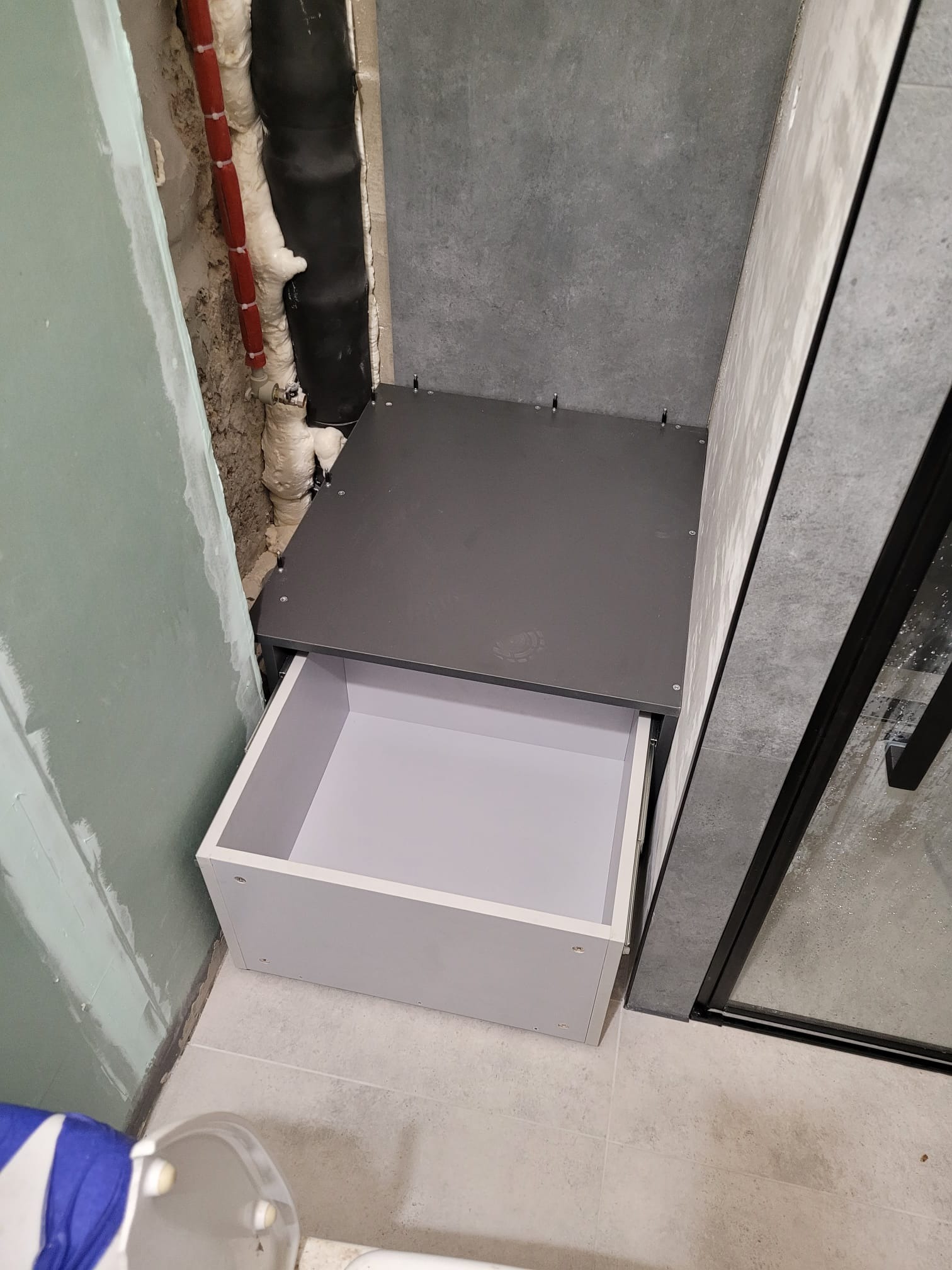 Wnęka z szufladą w łazience, widoczne rury z izolacją i fragment kabiny prysznicowej. Szuflada otwarta, biała. W tle ściana z szarych płytek.