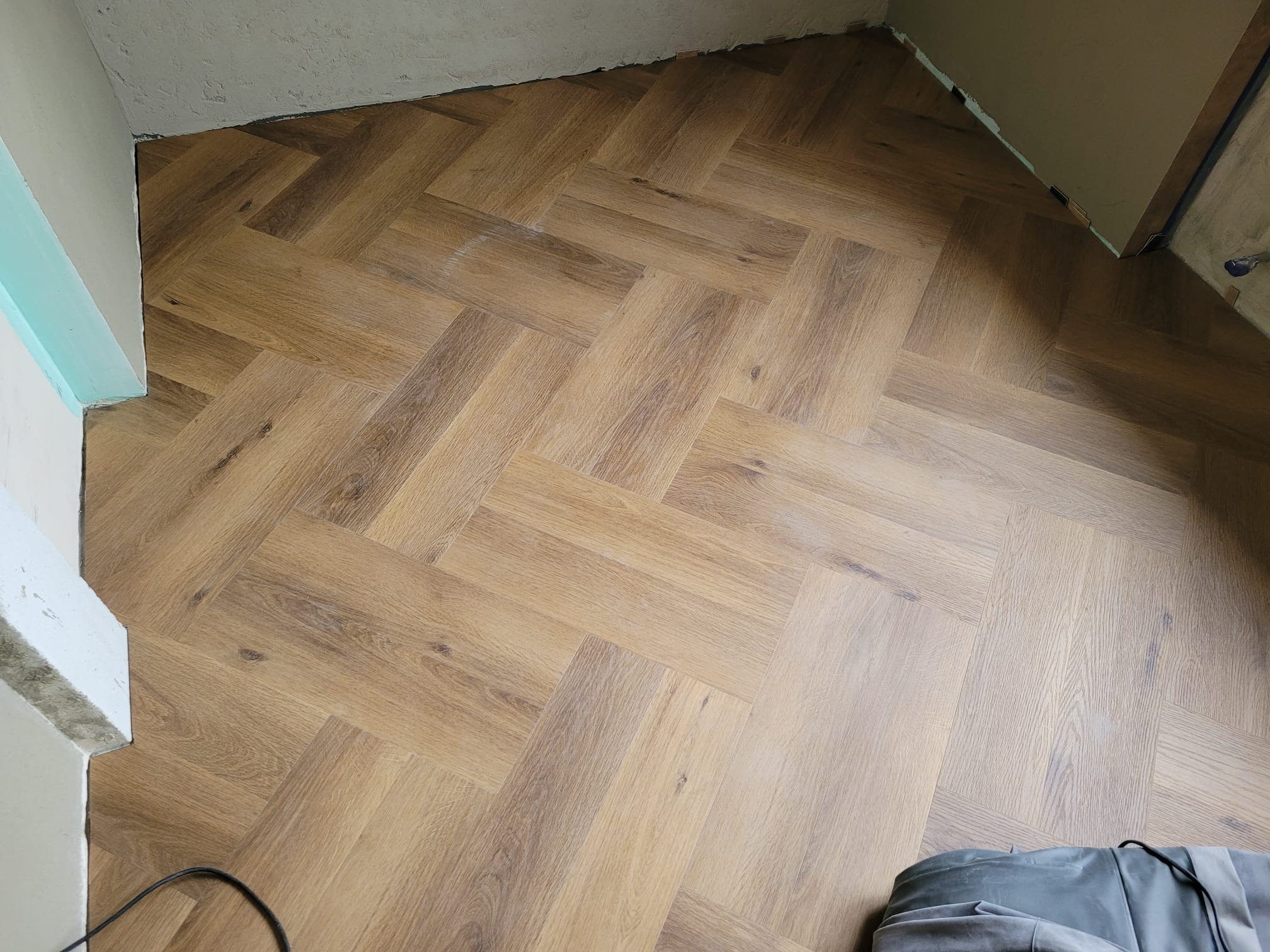 Ułożony parkiet w jodełkę, widoczne detale struktury drewna i fuga. Fragment ściany w tle. Podłoga po remoncie.