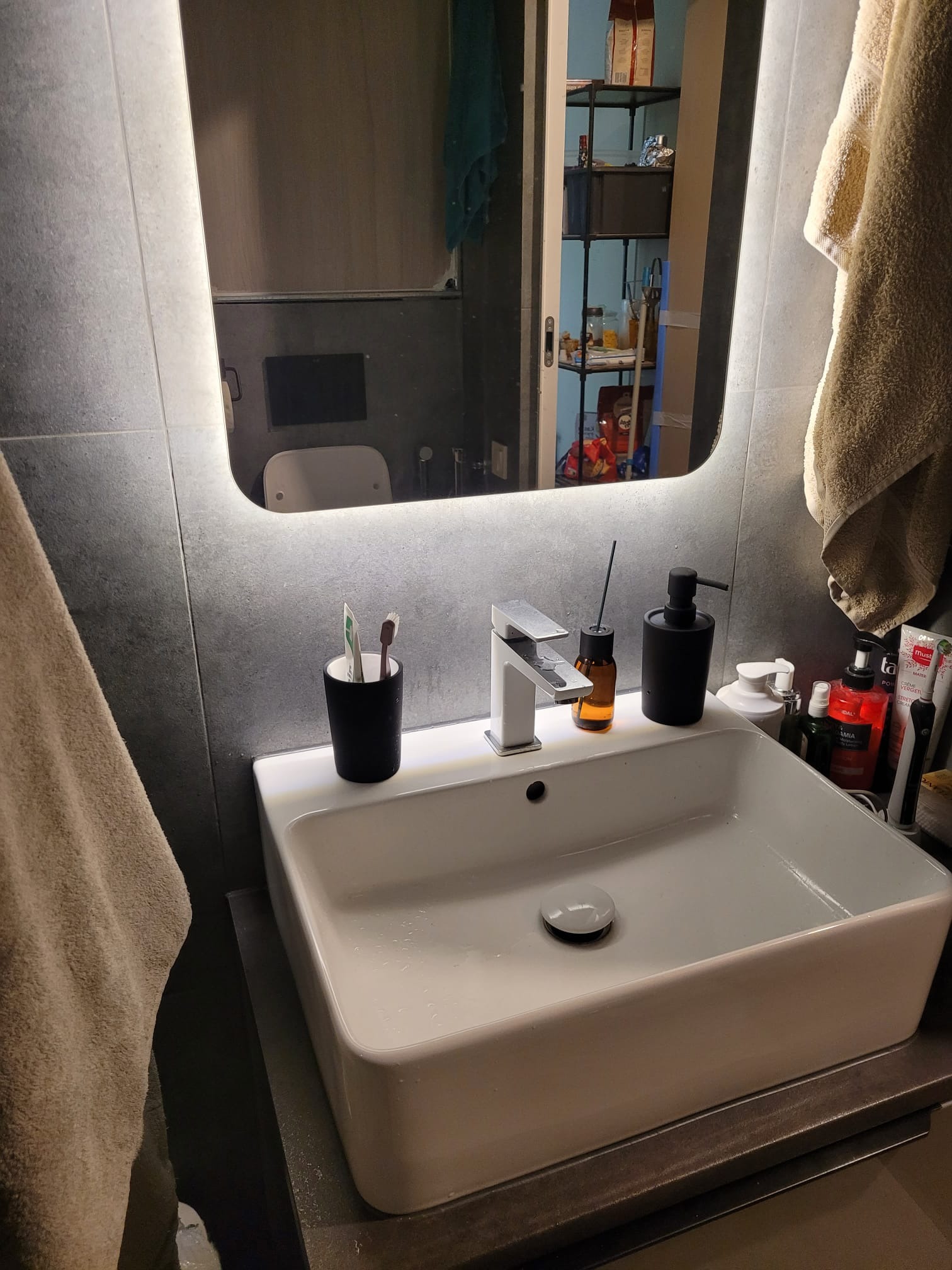 Biała, prostokątna umywalka z chromowaną baterią i akcesoriami łazienkowymi na blacie z szarego kamienia, odbicie w podświetlanym lustrze, fragment ręcznika.