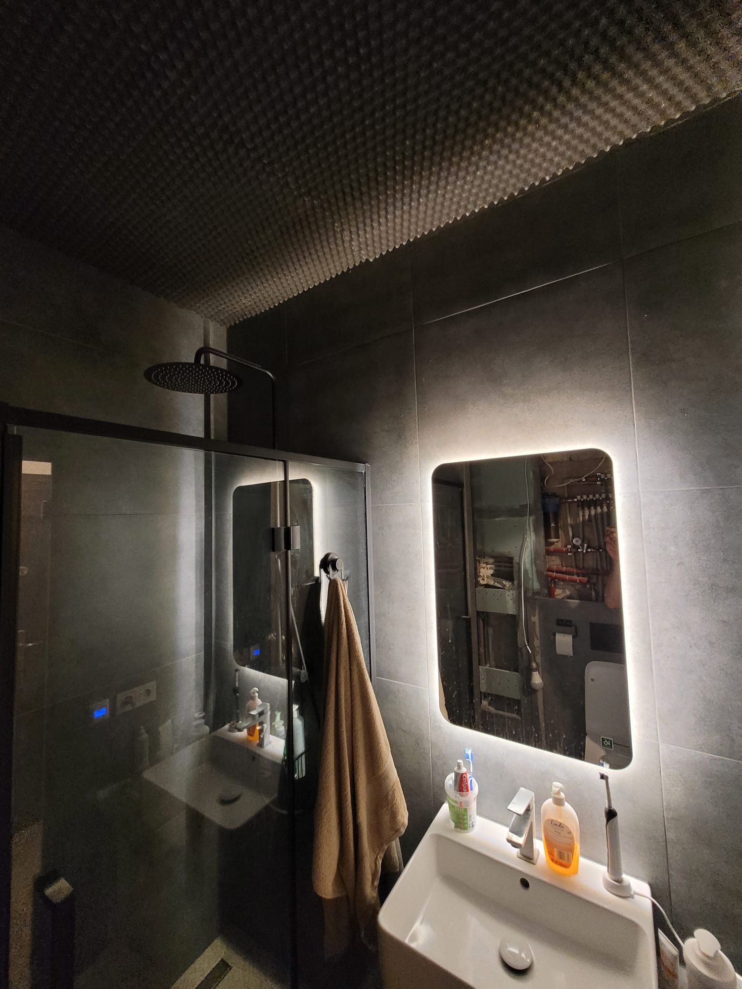 Nowoczesna łazienka z kabiną prysznicową i podświetlanym lustrem. Ciemne płytki, designerski sufit i armatura w kolorze czarnym tworzą elegancki klimat.