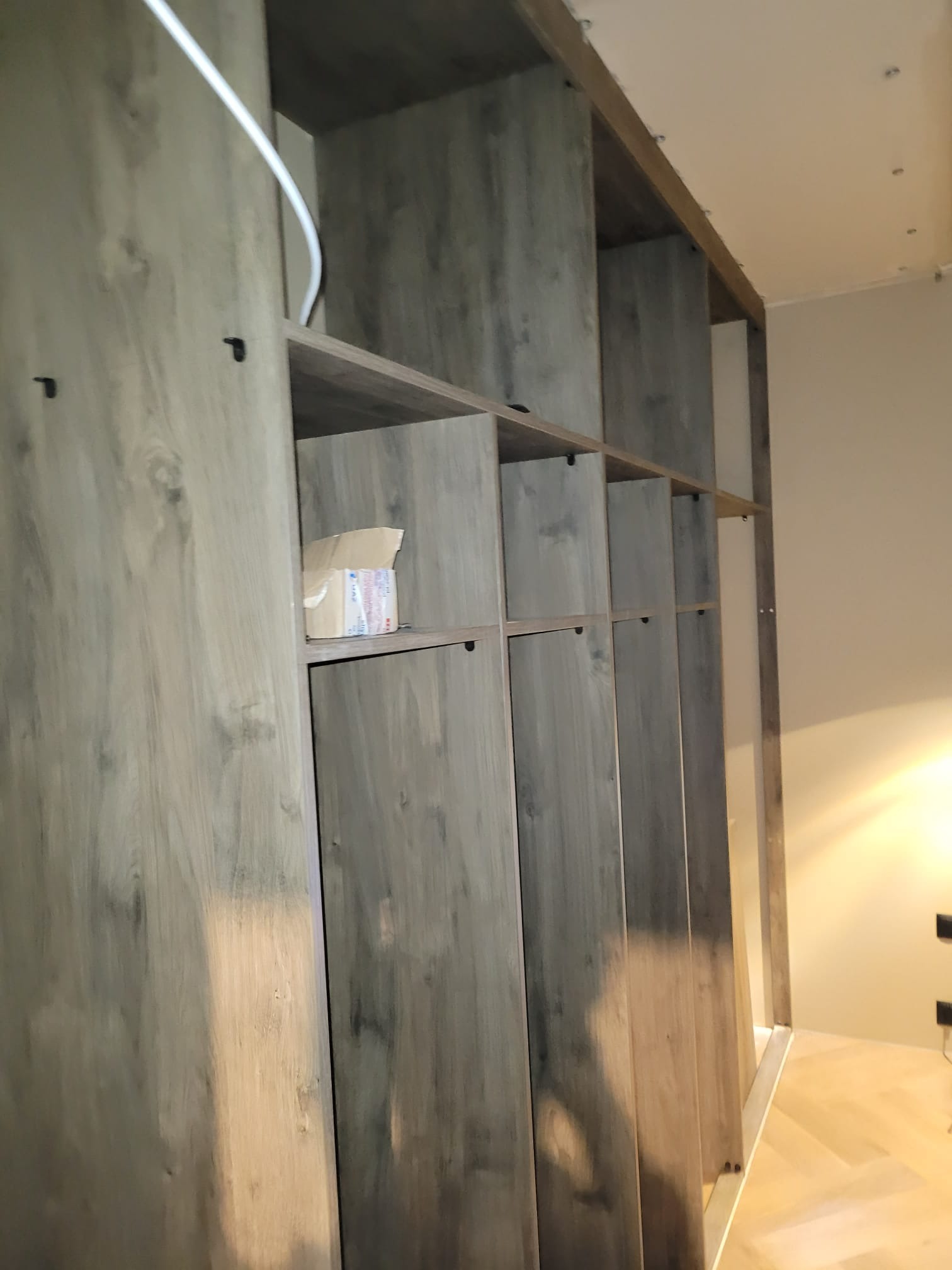 Drewniana szafa wnękowa z szarym, matowym wykończeniem, widoczne półki i drzwi. Na jednej z półek stoi karton. Widok z dołu, perspektywa podkreśla wysokość mebla.