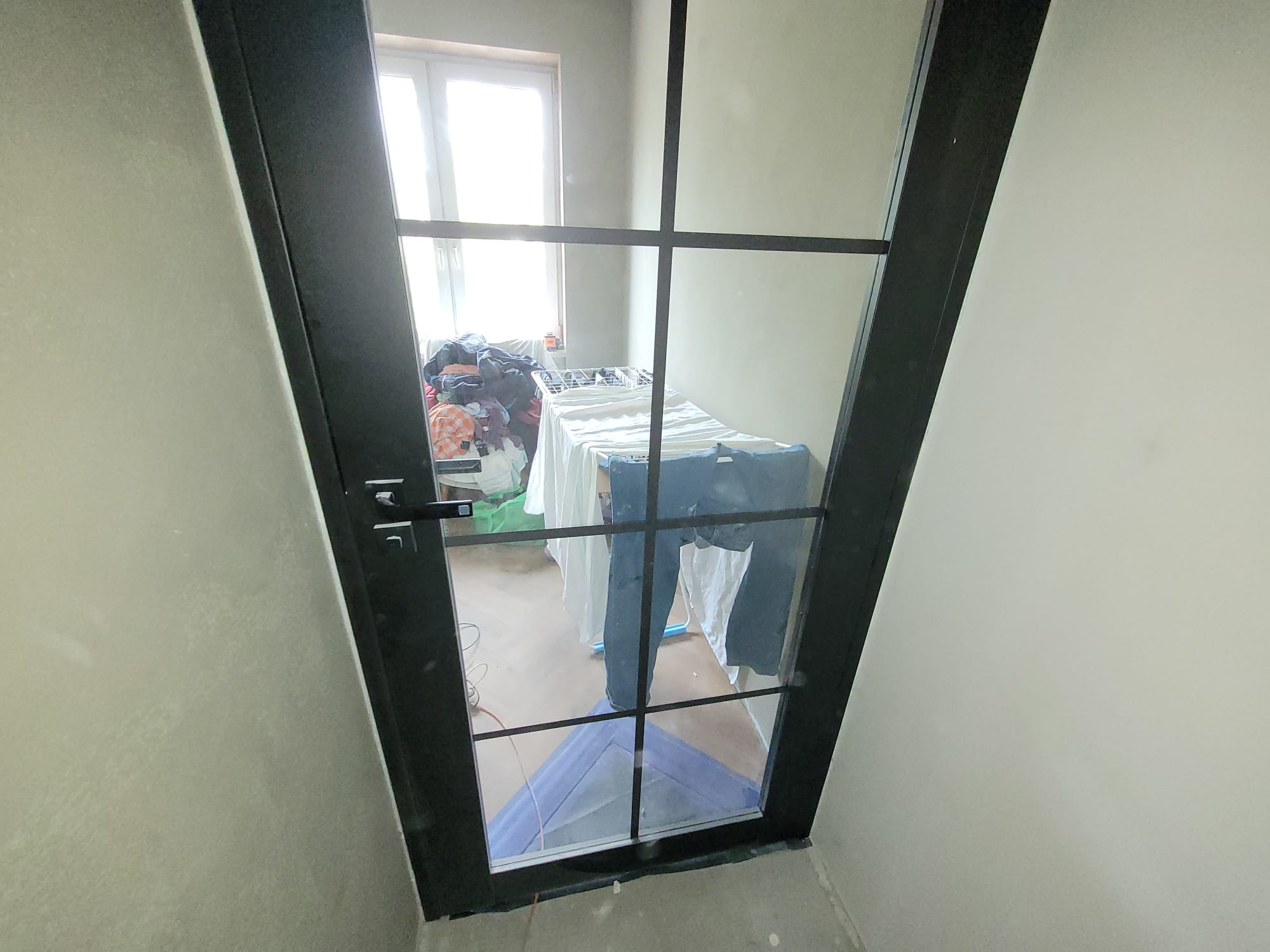 Ciemna, przeszklona rama drzwi z widokiem na suszarkę z praniem wewnątrz pomieszczenia. Surowy stan ścian, podłoga częściowo zabezpieczona folią.