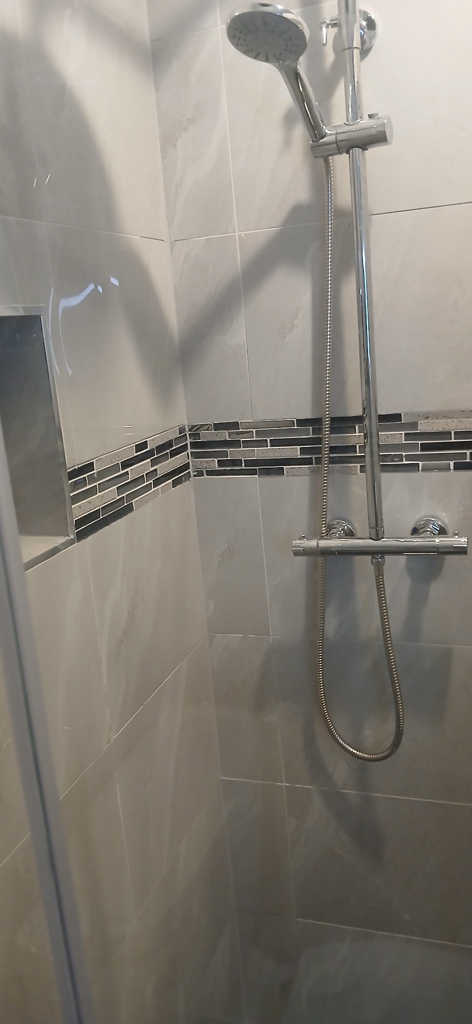 Wykończona kabina prysznicowa z szarymi płytkami i mozaiką. Chromowana armatura, deszczownica. Widok zza szklanej ściany kabiny.