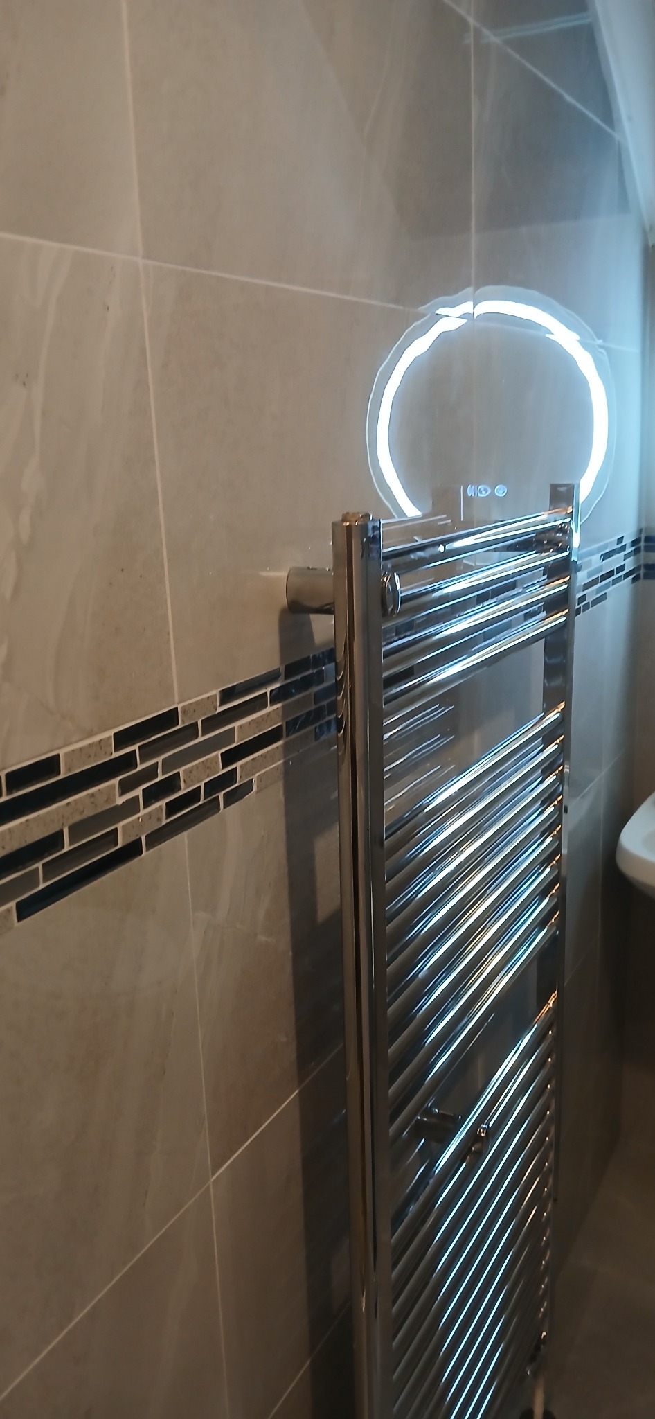 Nowoczesna łazienka z grzejnikiem drabinkowym i okrągłym lustrem LED. Szare płytki na ścianach z dekoracyjnym pasem mozaiki. Minimalistyczny design.