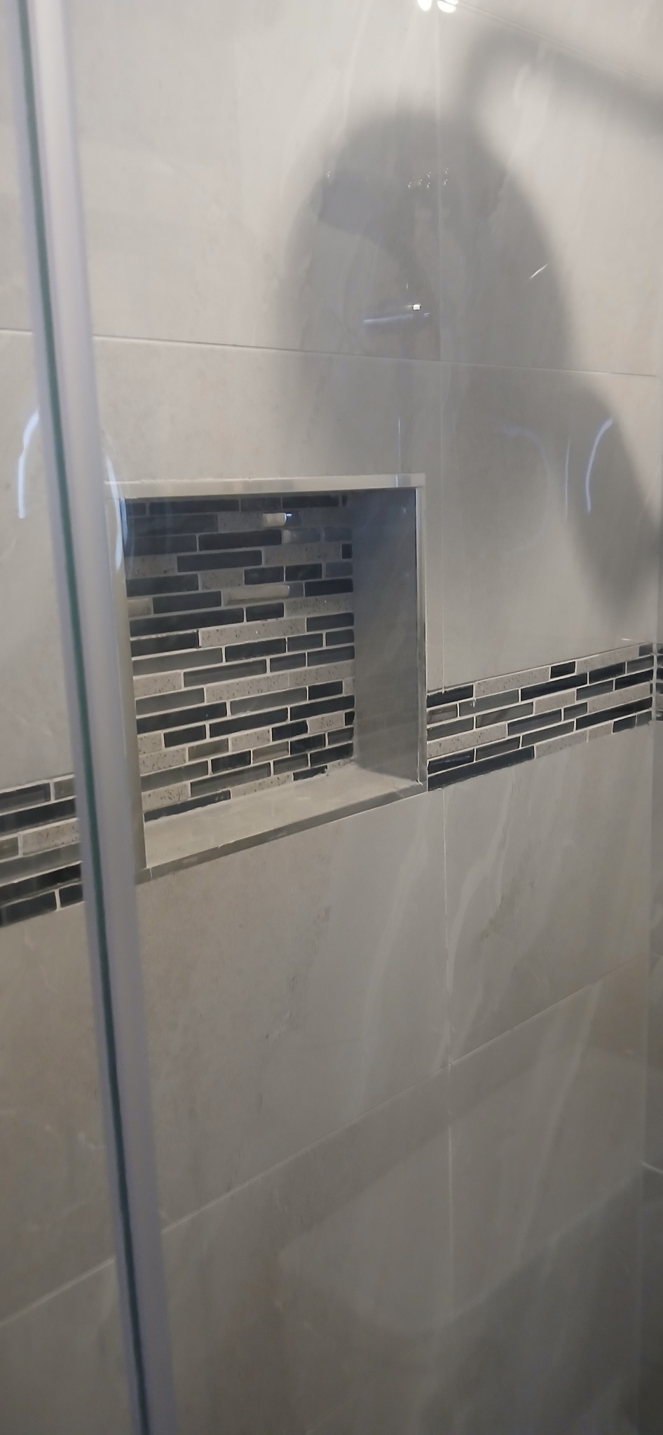 Wykończona wnęka z mozaiką w szarej łazience, widok przez szklaną kabinę. Minimalistyczny design, płytki imitujące marmur, poziome ułożenie mozaiki.