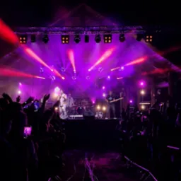 Zespół muzyczny na scenie oświetlonej fioletowymi światłami podczas koncertu plenerowego. Publiczność z podniesionymi rękami. Widoczne reflektory i elementy konstrukcji scenicznej.