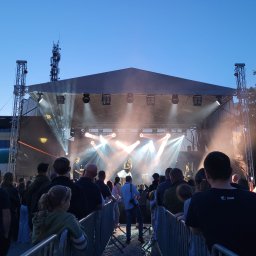 PMK Sound Mateusz Przytocki - Koncert plenerowy z zespołem na scenie oświetlonej reflektorami, widzowie obserwują występ. Scena zadaszona, widoczne elementy konstrukcji i oświetlenia.