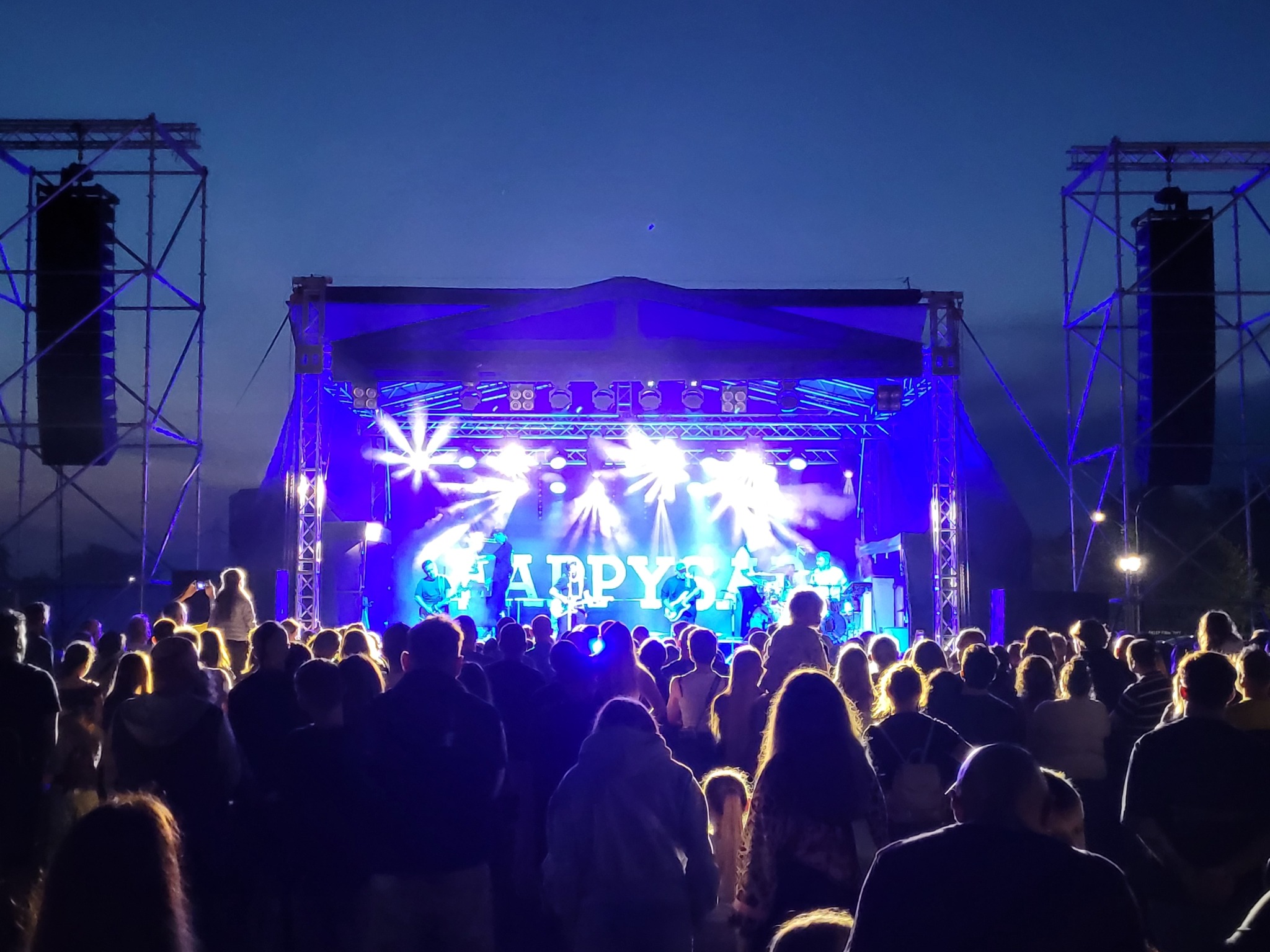 Koncert plenerowy z oświetloną sceną, na której występuje zespół 'HAPPY SAD', widoczny tłum publiczności, wieczorny klimat.