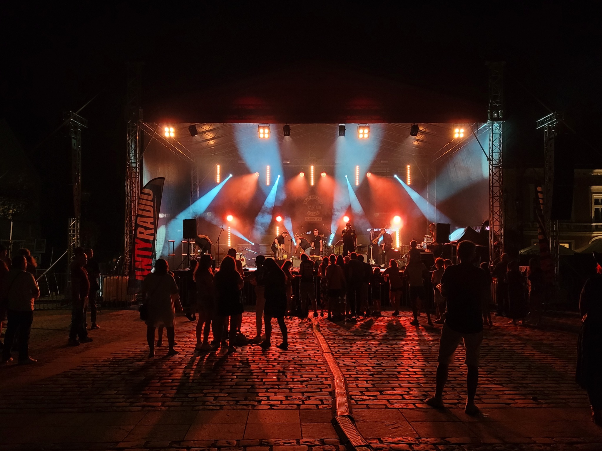 Scena z oświetleniem eventowym podczas koncertu plenerowego, widzowie w tle. Czerwono-niebieskie światła, flaga z napisem 'MyRadio' po lewej stronie.