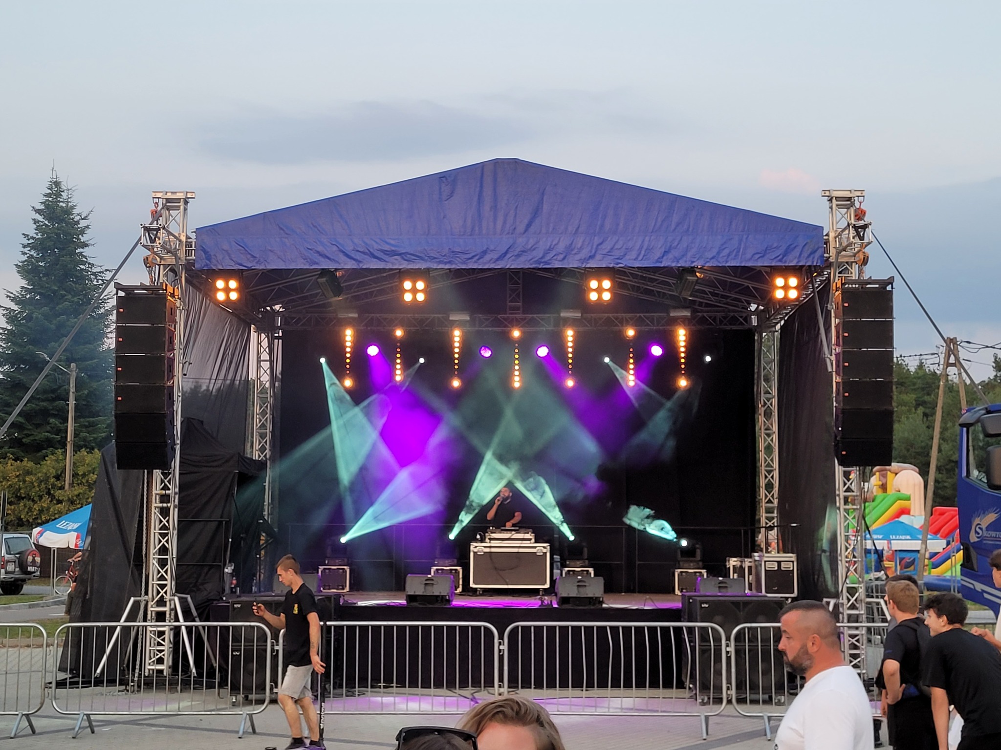 Scena z nagłośnieniem i oświetleniem podczas imprezy plenerowej, widoczny DJ za konsoletą, efekty świetlne, publiczność w tle i elementy konstrukcji scenicznej.