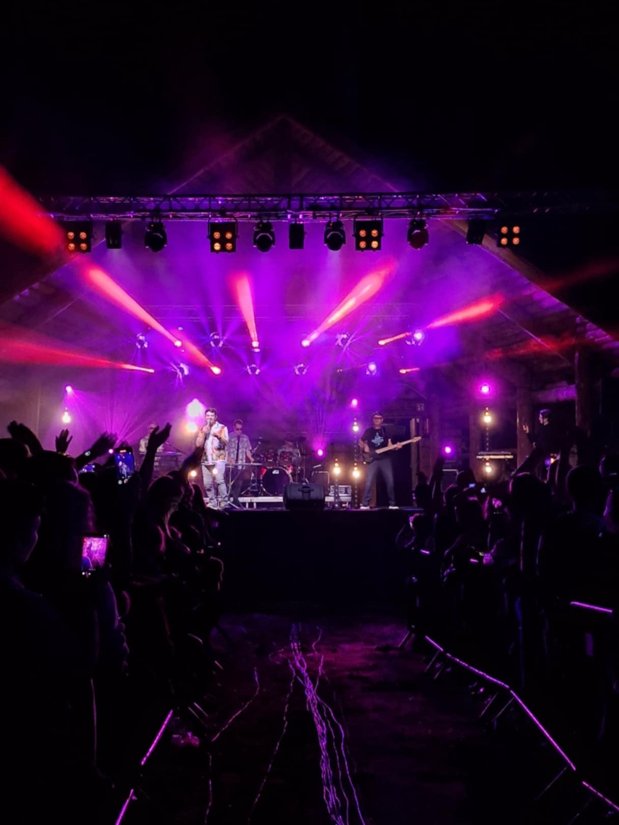 Zespół muzyczny na scenie oświetlonej fioletowymi światłami podczas koncertu plenerowego. Publiczność z podniesionymi rękami. Widoczne reflektory i elementy konstrukcji scenicznej.