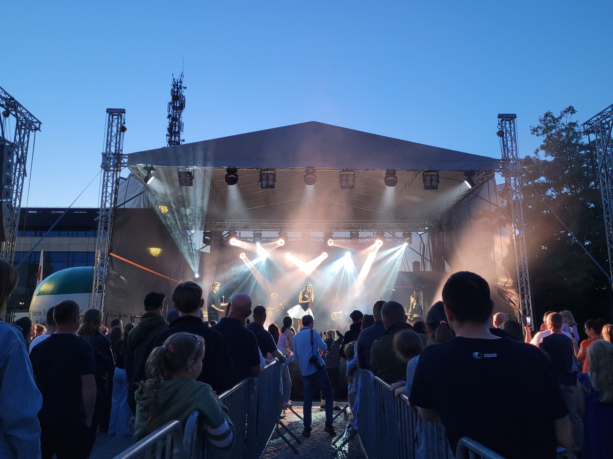 Koncert plenerowy z zespołem na scenie oświetlonej reflektorami, widzowie obserwują występ. Scena zadaszona, widoczne elementy konstrukcji i oświetlenia.