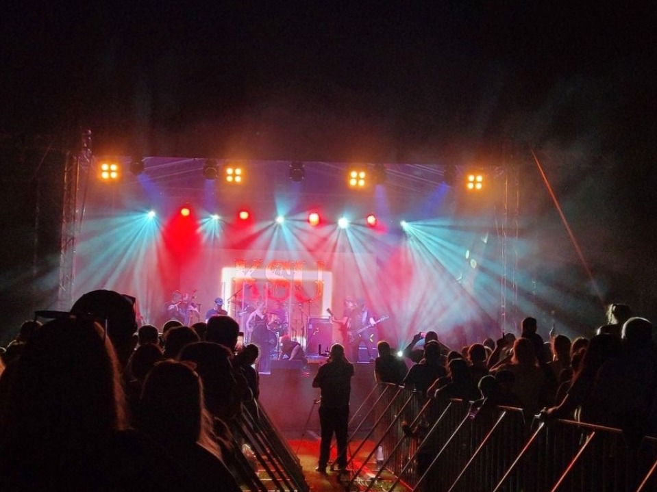 Koncert plenerowy z widokiem na scenę oświetloną czerwonymi i niebieskimi reflektorami, tłum ludzi obserwuje występ zespołu na żywo, widoczne barierki zabezpieczające.