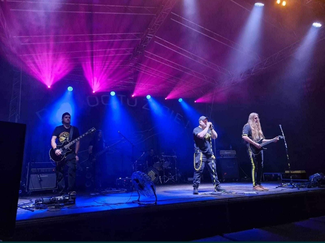 Zespół muzyczny na scenie podczas koncertu. Wokalista śpiewa do mikrofonu, gitarzyści grają na gitarach, oświetlenie sceniczne w kolorach niebieskim i różowym.