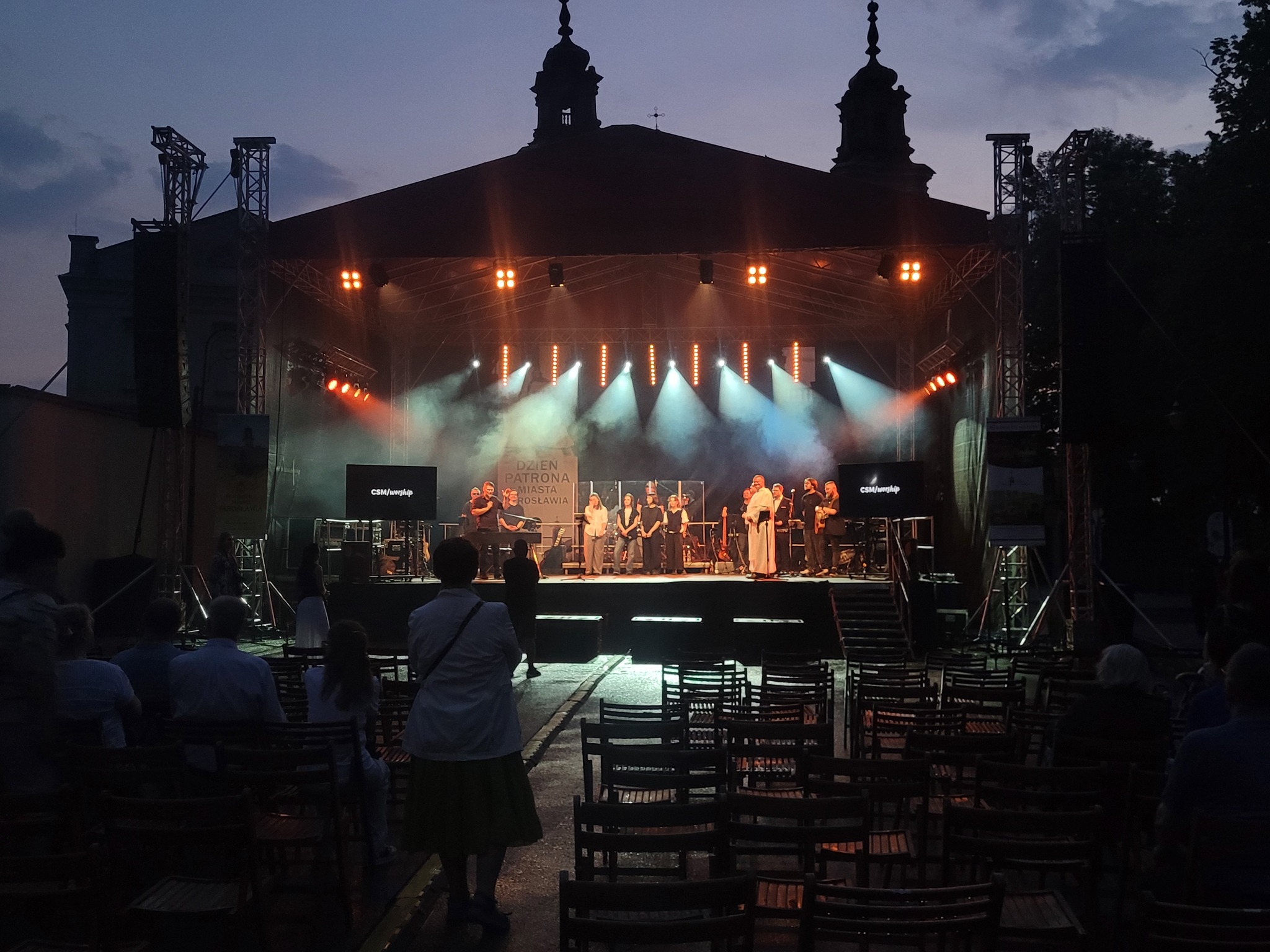 Scena plenerowa z oświetleniem i zespołem muzycznym podczas wieczornego wydarzenia. Widzowie siedzą na krzesłach, w tle budynek z wieżami i napis 'Dzień Patrona'.