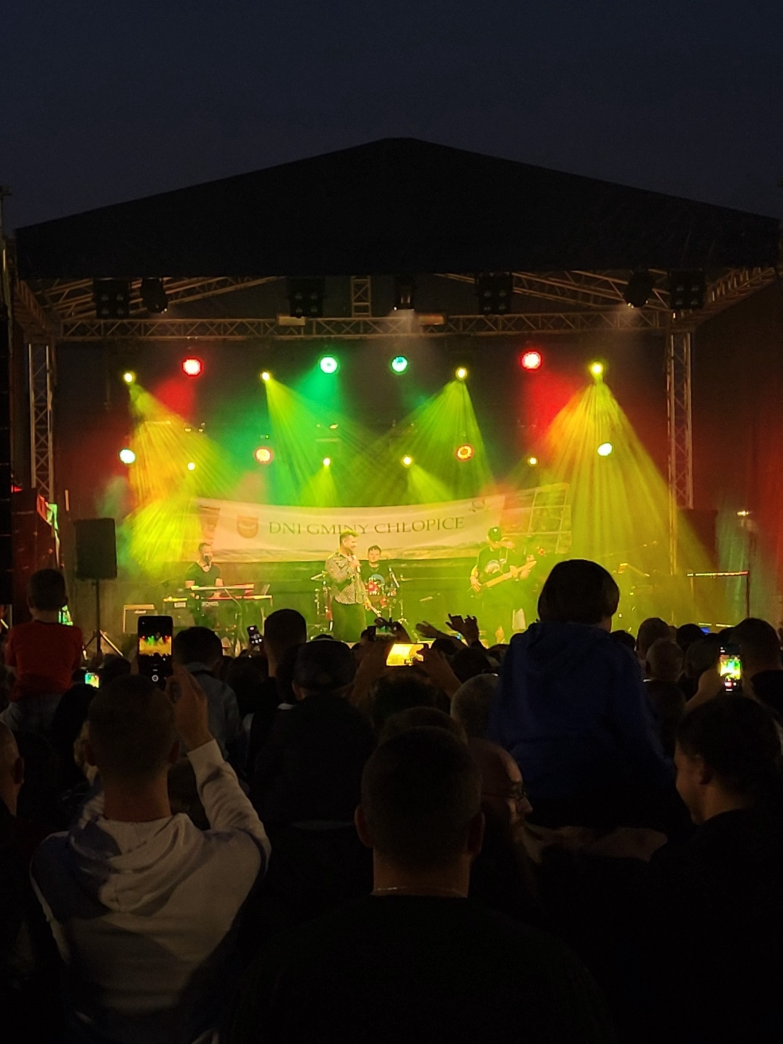 Zespół muzyczny na scenie podczas plenerowego festiwalu. Widzowie filmują koncert telefonami. Kolorowe oświetlenie sceniczne tworzy dynamiczny efekt.