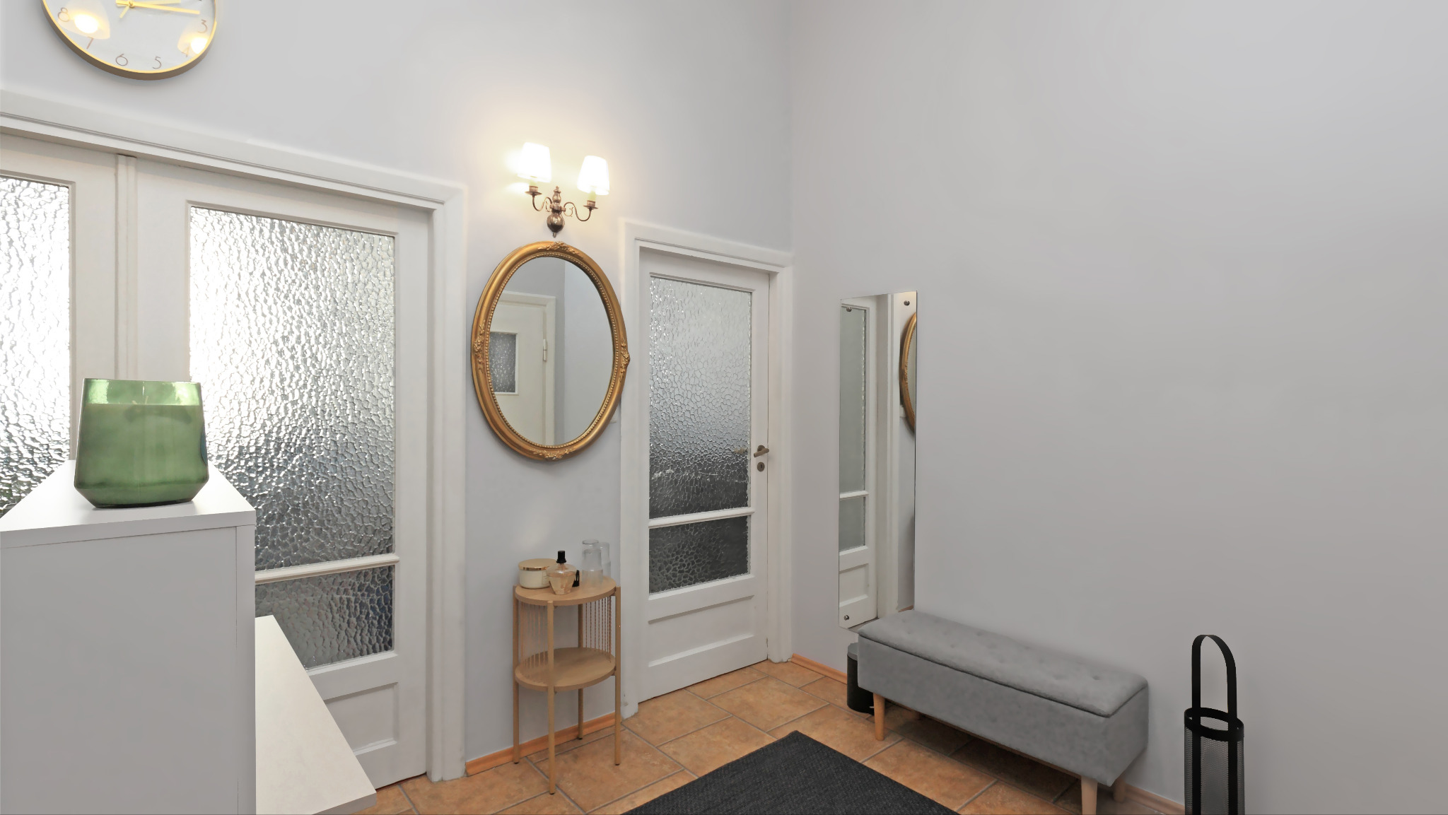 Jasne wnętrze przedpokoju z lustrami, białymi drzwiami z falistym szkłem i elementami dekoracyjnymi w kolorach złota i szarości. Uwagę przykuwa zegar na ścianie.