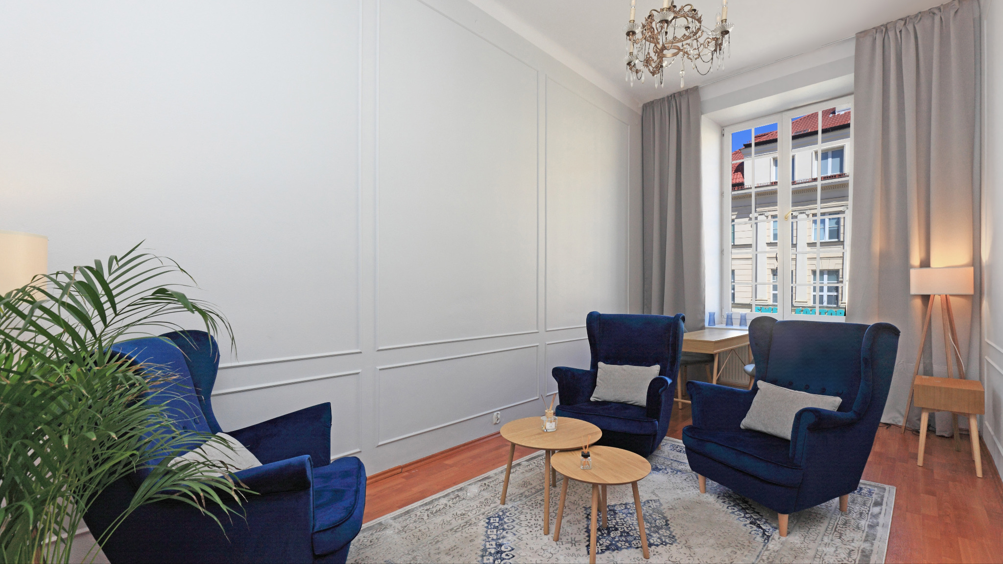 Jasne wnętrze gabinetu z trzema granatowymi fotelami, stolikami kawowymi, rośliną i oknem z widokiem na budynek. Spokojna aranżacja przestrzeni.