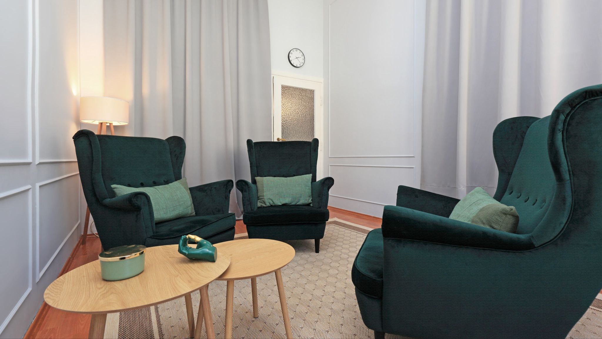 Klimatyczne wnętrze gabinetu z trzema fotelami w kolorze butelkowej zieleni, drewnianymi stolikami i stonowaną kolorystyką ścian. Przytulna przestrzeń dla pacjentów.