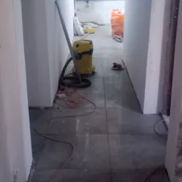 Korytarz w trakcie prac wykończeniowych: świeżo położone ciemne płytki, odkurzacz przemysłowy, widoczne instalacje pod sufitem. W tle materiały budowlane.