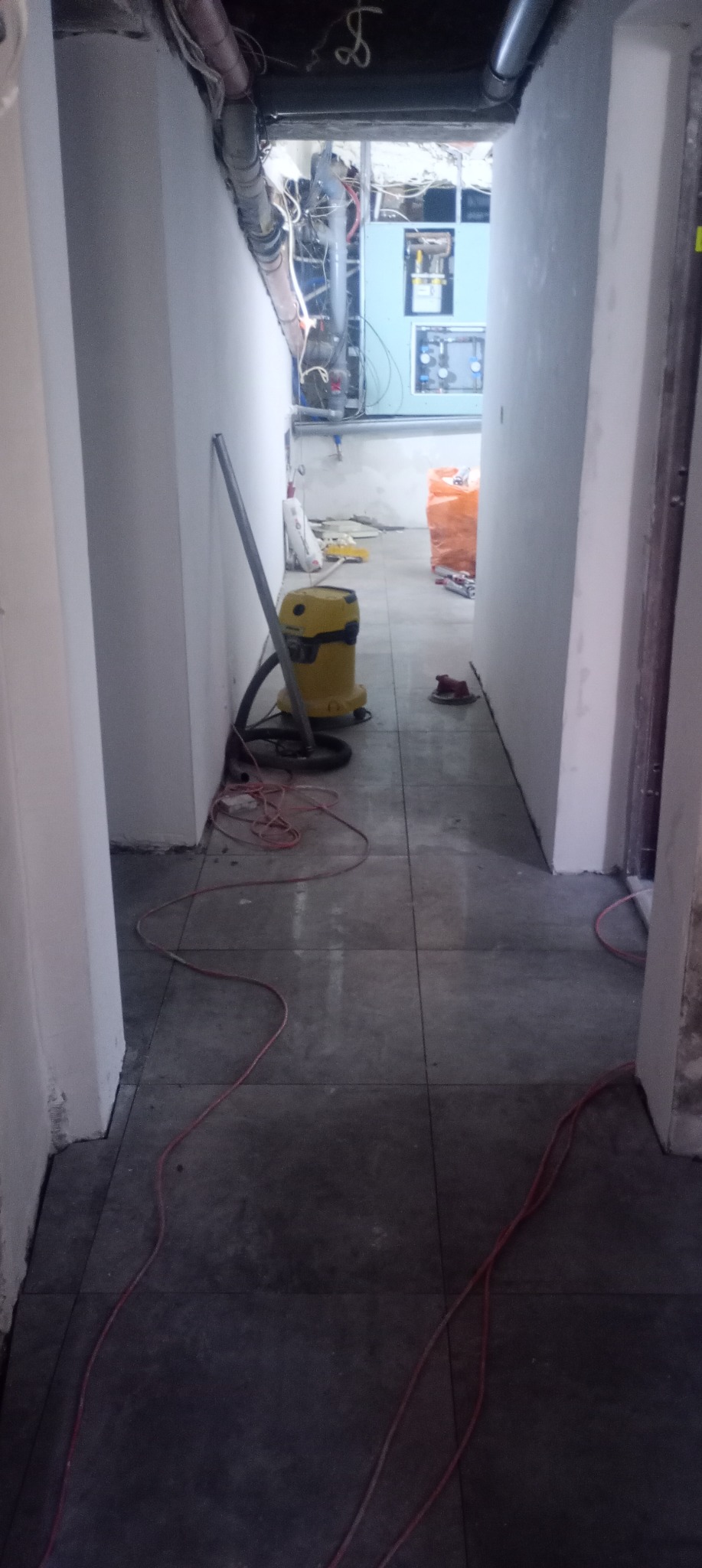 Korytarz w trakcie prac wykończeniowych: świeżo położone ciemne płytki, odkurzacz przemysłowy, widoczne instalacje pod sufitem. W tle materiały budowlane.