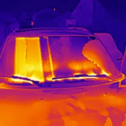 Termowizyjne zdjęcie samochodu z otwartymi drzwiami, ukazujące rozkład temperatury na szybie przedniej i karoserii, wykonane w odcieniach pomarańczy i fioletu.