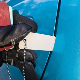 MICHAŁ KOWALÓWKA M&M BROKERS - Błękitny lakier auta sprawdzany przez rzeczoznawcę w czarnych rękawicach, z użyciem próbnika lakieru na łańcuszku. Kontrola grubości powłoki.