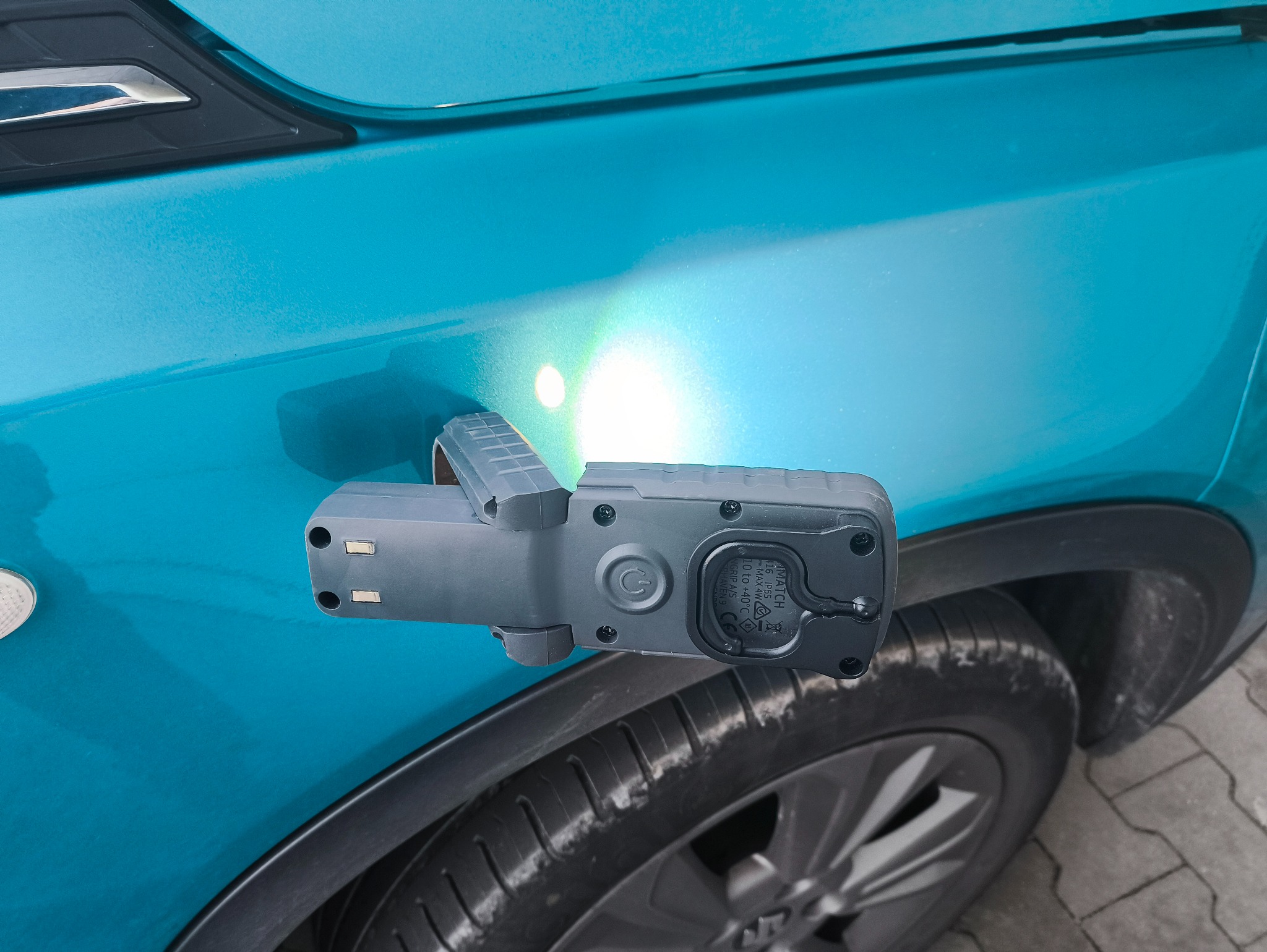 Lampa inspekcyjna przylegająca do karoserii turkusowego auta, oświetlająca lakier. Widoczne koło i fragment nadkola. Detailing samochodowy w trakcie pracy.