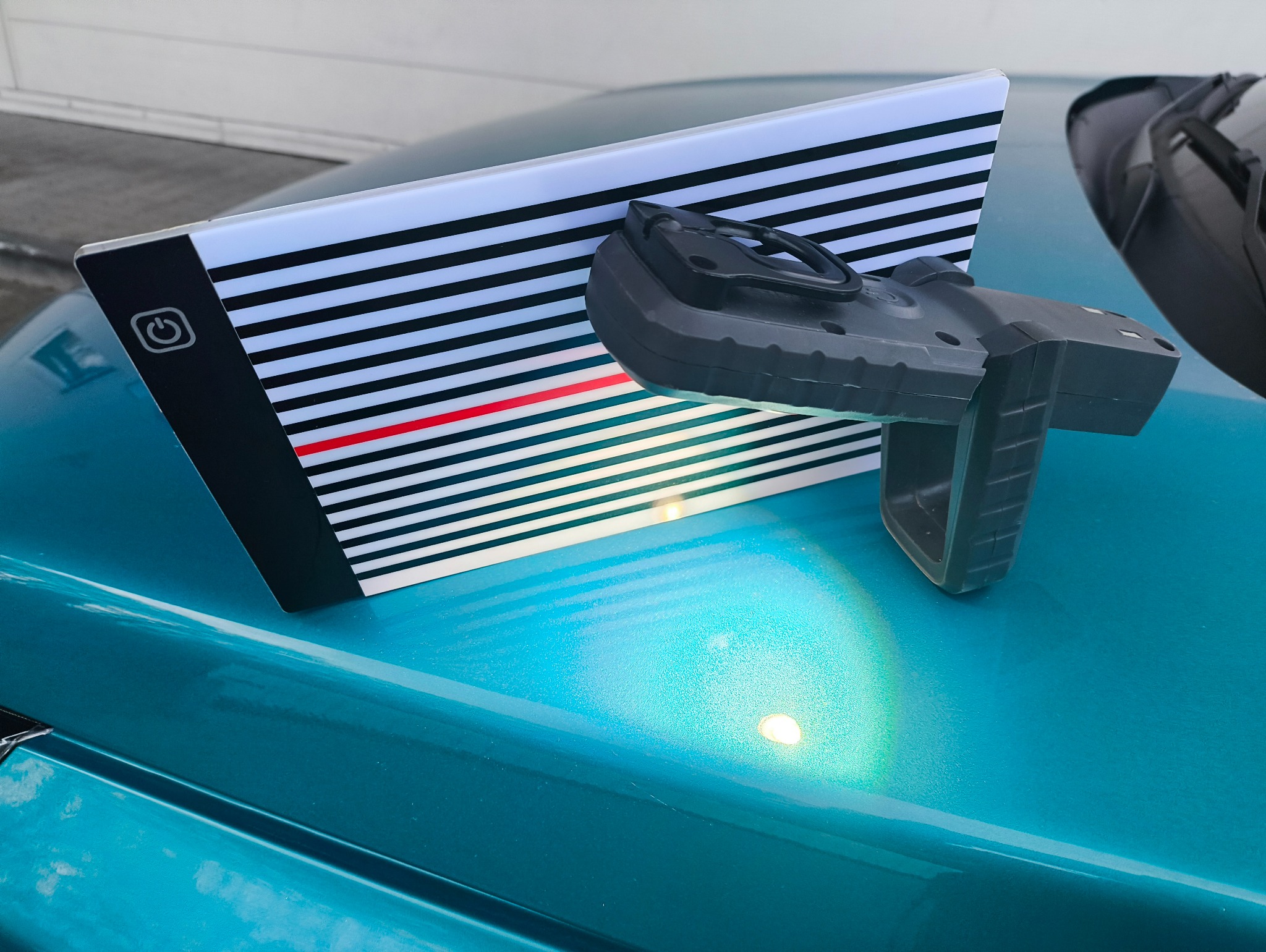 Turkusowy lakier samochodu z panelem inspekcyjnym w czarno-białe paski i lampą do wykrywania wgnieceń. Narzędzia do oceny uszkodzeń karoserii.