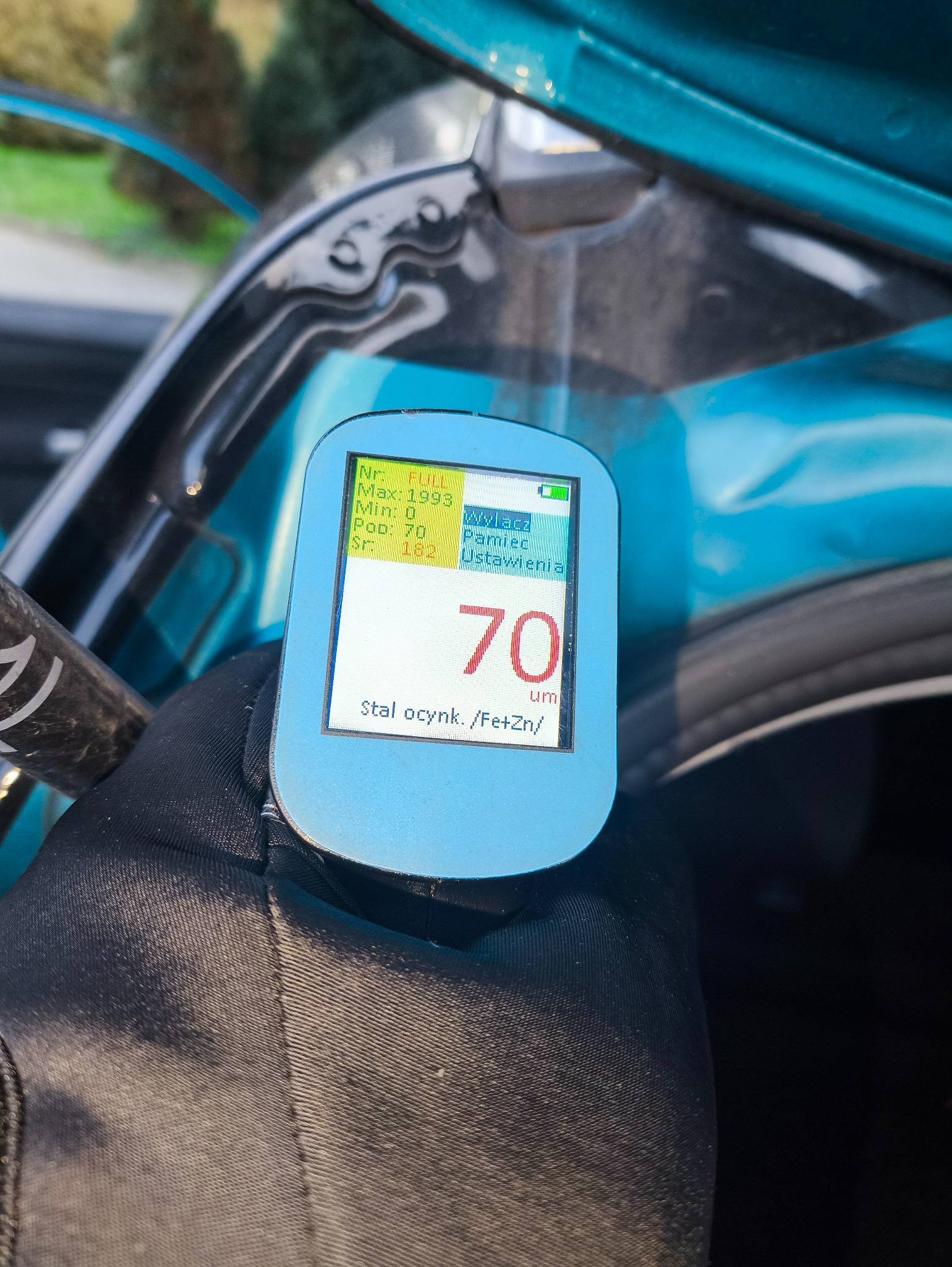 Pomiar grubości lakieru na aucie miernikiem z wyświetlaczem wskazującym 70 um, stal ocynkowana. Widoczna ręka w czarnej rękawicy trzymająca urządzenie.