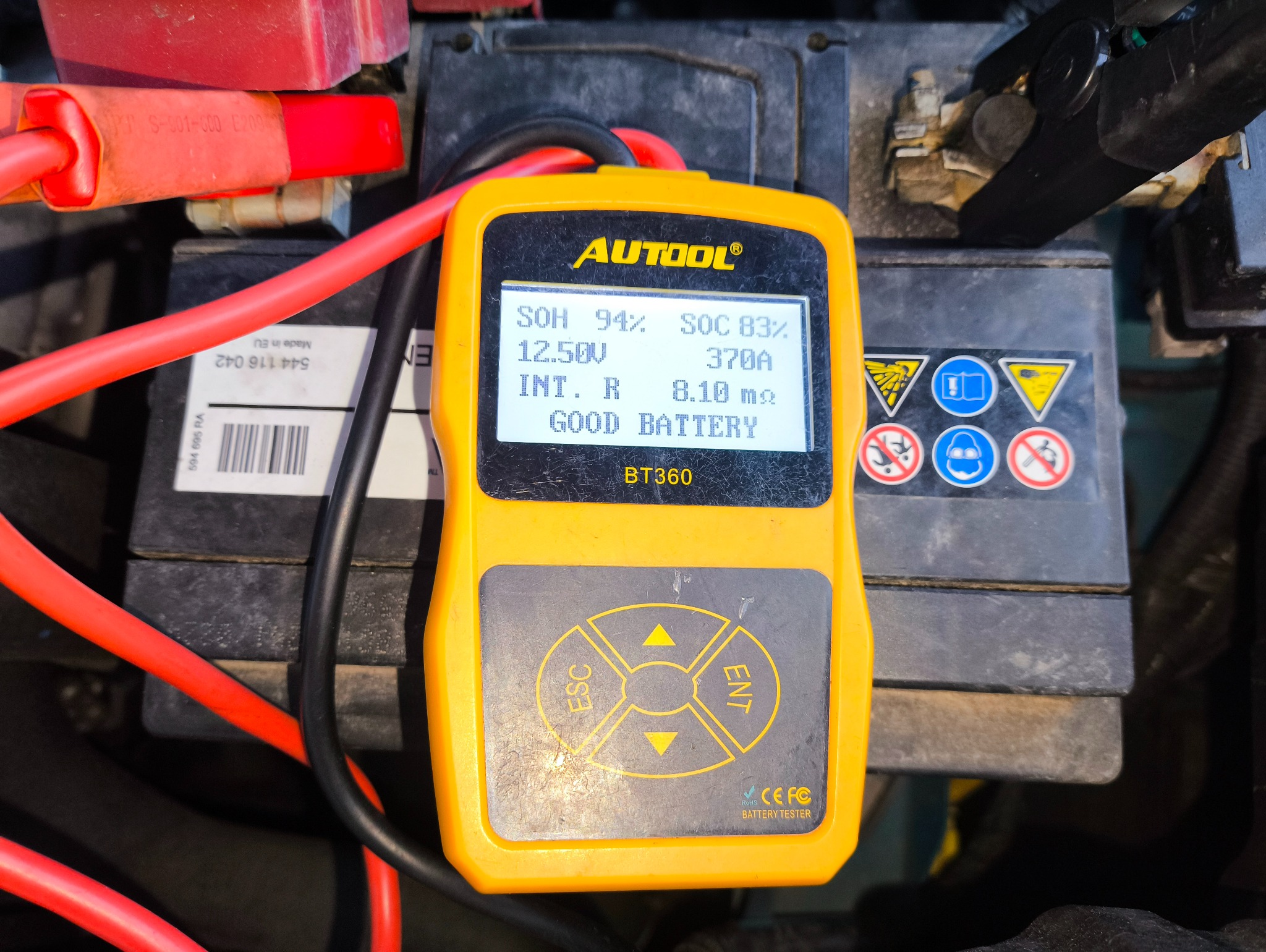 Tester akumulatora Autool BT360 wskazuje 'Good Battery' na tle akumulatora samochodowego z czerwonymi klemami. Widoczne parametry: SOH 94%, SOC 83%, 12.50V.