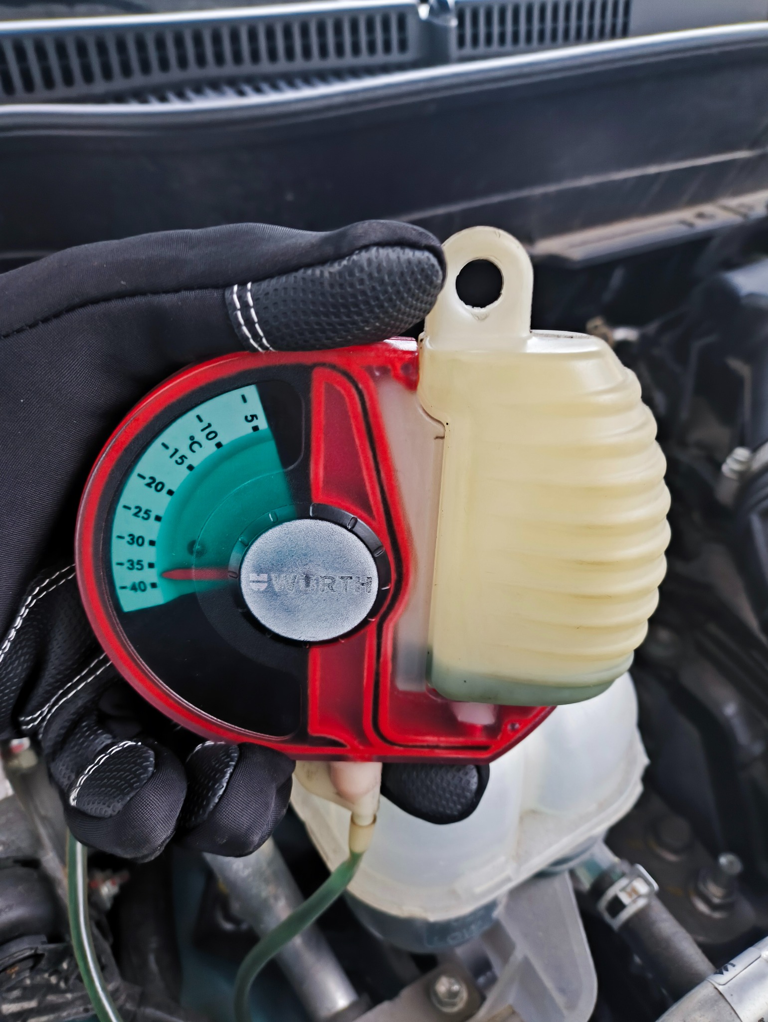 Pomiar temperatury płynu chłodniczego w samochodzie za pomocą refraktometru marki Wurth, trzymanego w rękawicy ochronnej na tle silnika.