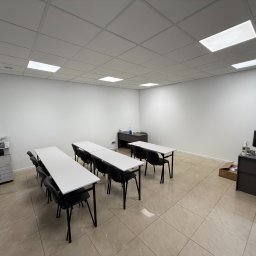 EL-GROUP MAŁGORZATA KUBIEŃ, SEBASTIAN GOŁĄB SPÓŁKA CYWILNA - Jasna sala szkoleniowa z rzędami białych stołów i czarnych krzeseł, biurko z komputerem, drukarka Canon w folii i kasetonowe oświetlenie sufitowe.