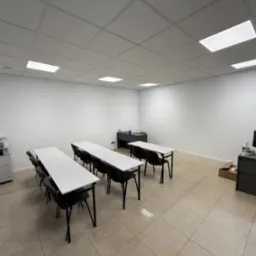 Jasna sala szkoleniowa z rzędami białych stołów i czarnych krzeseł, biurko z komputerem, drukarka Canon w folii i kasetonowe oświetlenie sufitowe.