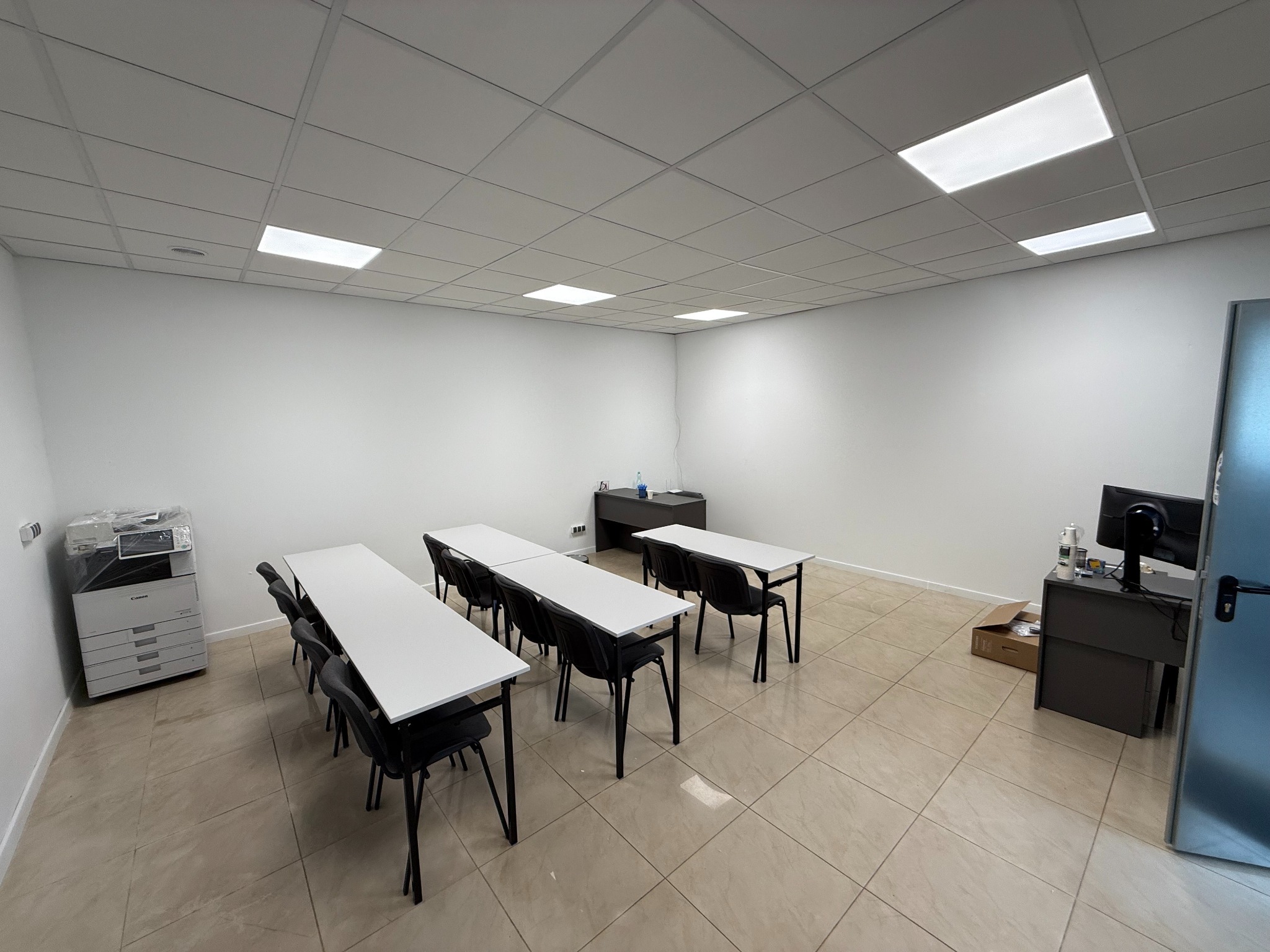 Jasna sala szkoleniowa z rzędami białych stołów i czarnych krzeseł, biurko z komputerem, drukarka Canon w folii i kasetonowe oświetlenie sufitowe.