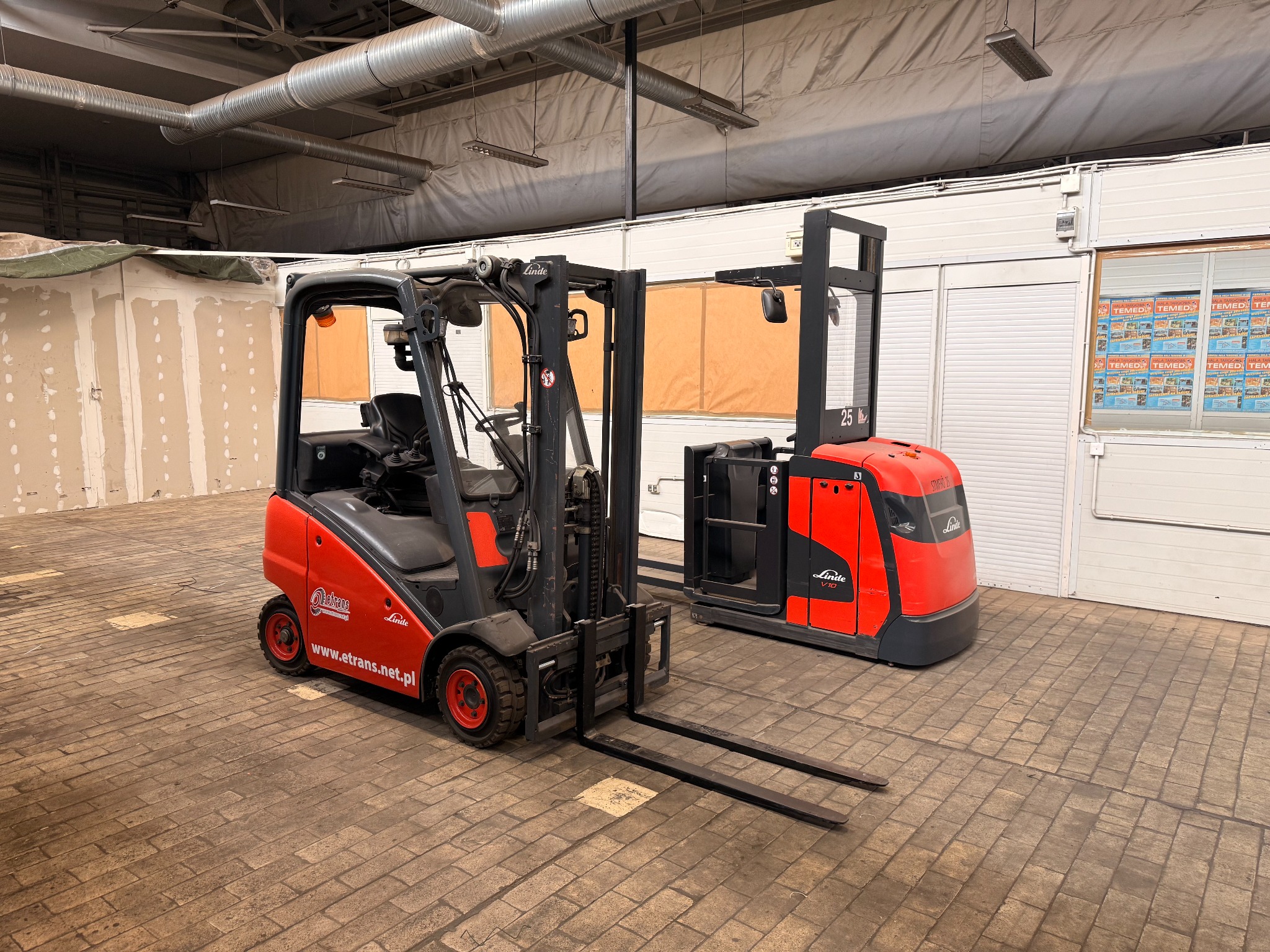 Dwa czerwono-czarne wózki widłowe Linde, jeden z widłami, drugi platformowy, stoją na hali z betonową podłogą. Na wózku widoczny adres etrans.net.pl.