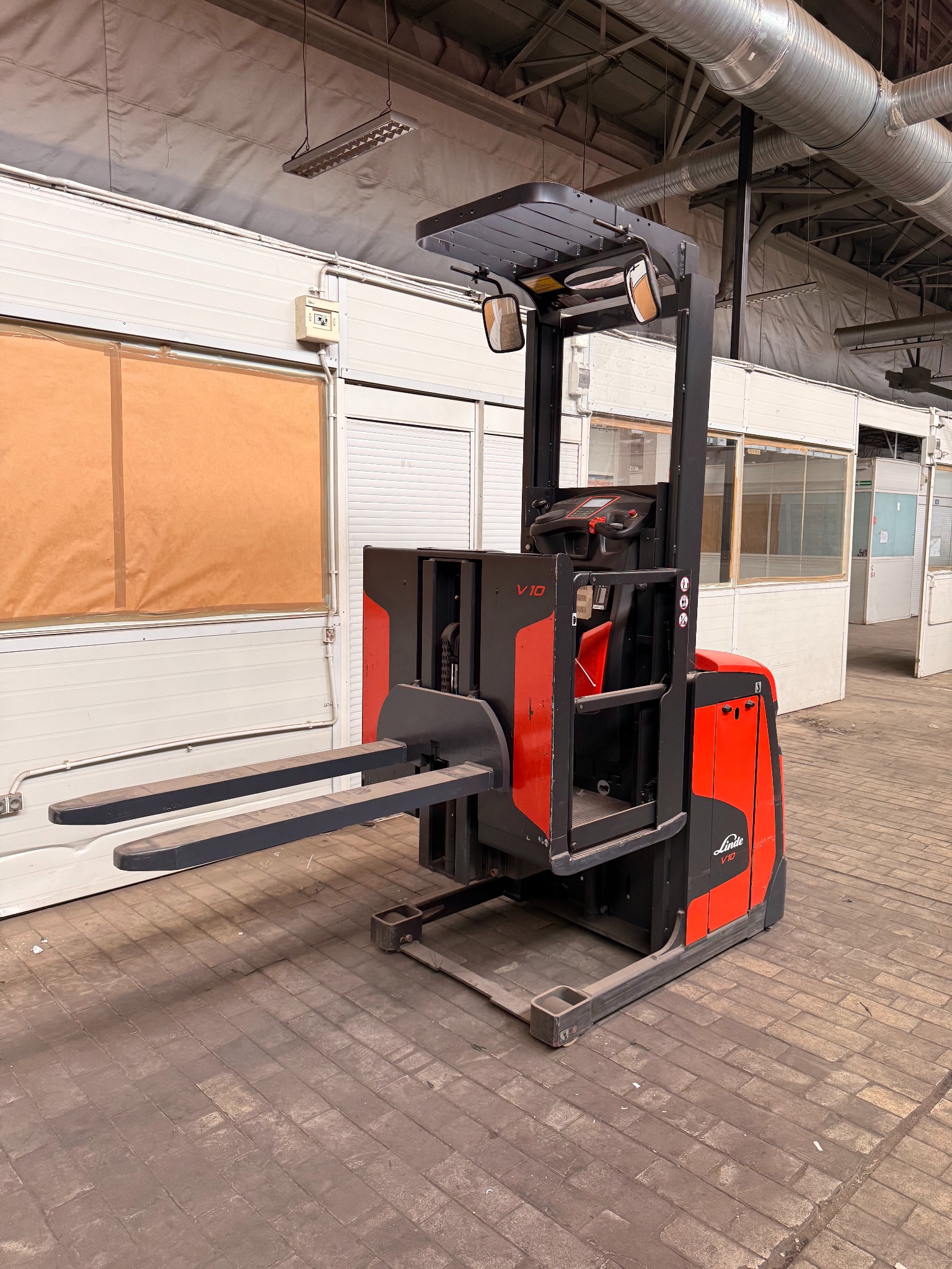 Czerwono-czarny wózek widłowy Linde V10 na tle magazynu z widocznymi instalacjami na suficie i ścianami z oknami. Podłoże z cegieł.