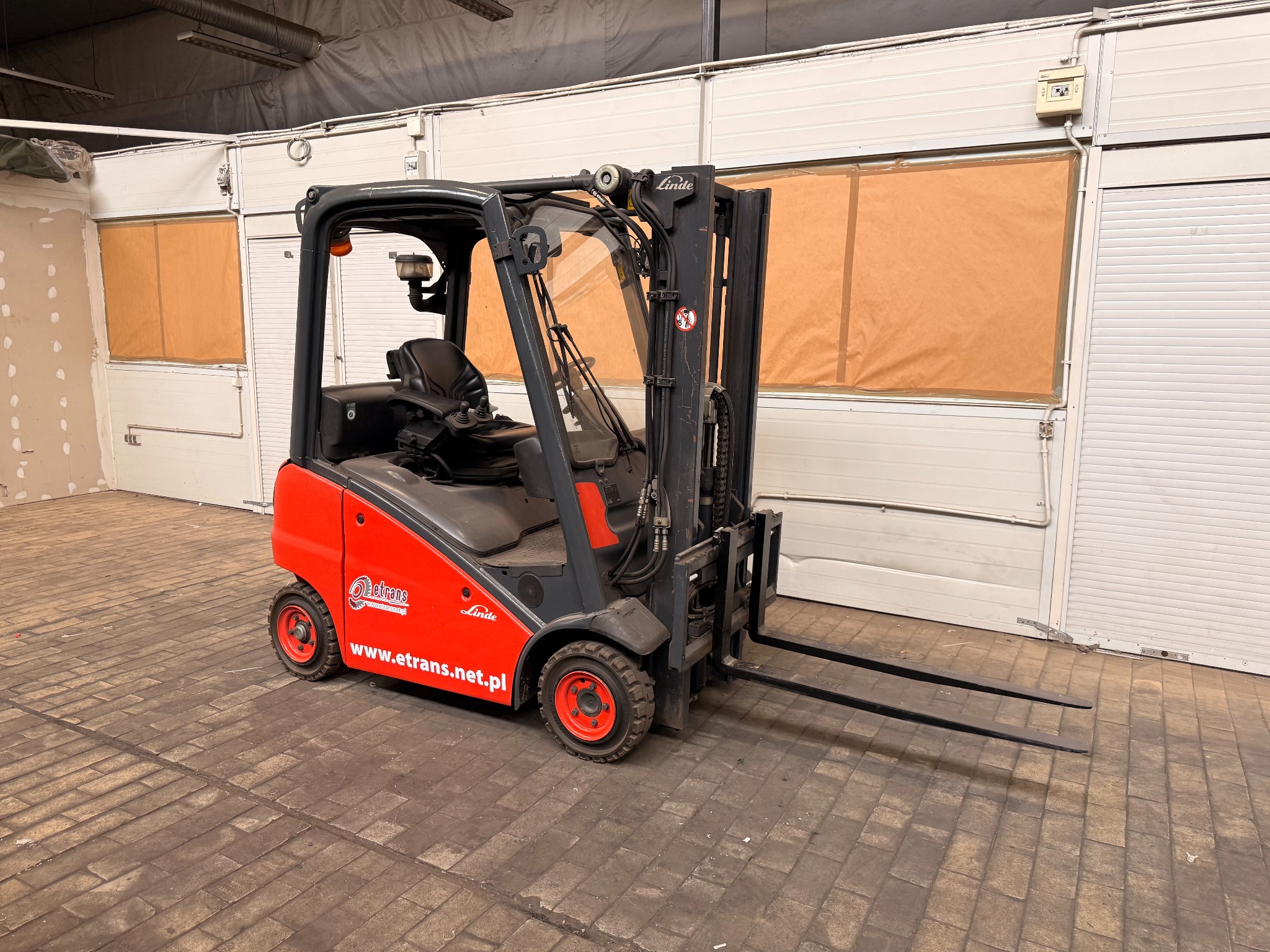 Czerwono-szary wózek widłowy Linde z otwartą kabiną na tle magazynu z oknami zabezpieczonymi brązowym papierem. Na wózku widoczne logo firmy etrans.net.pl.