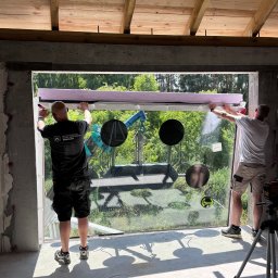 SAMPRE Bydgoszcz - Łukasz Rabęda - Dwóch pracowników montuje dużą, panoramiczną szybę w surowym otworze betonowego budynku, używając specjalistycznego sprzętu widocznego na zewnątrz. Wnętrze z drewnianym stropem.
