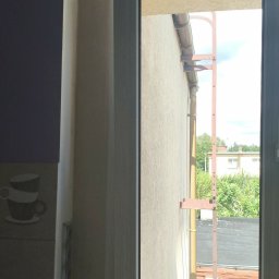 SAMPRE Bydgoszcz - Łukasz Rabęda - Nowoczesne okno z jasną ramą, z widokiem na elewację budynku z rynną i metalową konstrukcją. Wnętrze z ciemną ścianą i dekoracyjnymi płytkami z filiżankami.