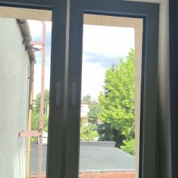 SAMPRE Bydgoszcz - Łukasz Rabęda - Dwuskrzydłowe okno z jasnoszarą ramą, zamontowane w ciemnej ścianie, z widocznym uszczelnieniem. Zewnętrzny widok na zielone drzewa, płaski dach i rynnę budynku.