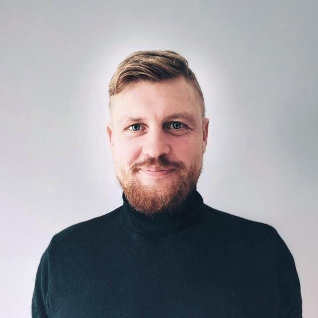 Portret mężczyzny z rudą brodą, ubranego w czarny golf, na jasnoszarym tle. Uśmiechnięty, z niebieskimi oczami i modną fryzurą.