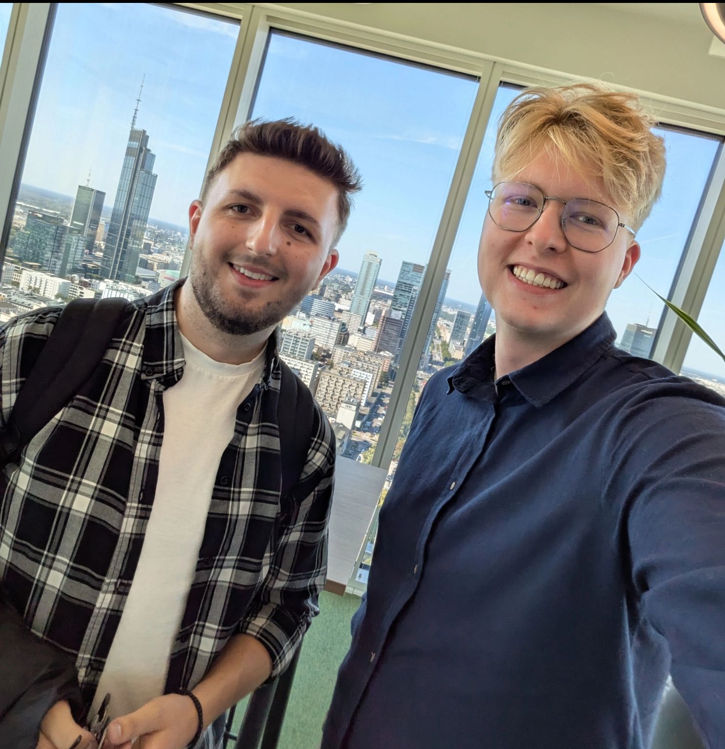 Uśmiechnięci mężczyźni na tle panoramy Warszawy z wieżowcami, prezentujący nowoczesne biuro doradcy finansowego lub brokera ubezpieczeniowego.