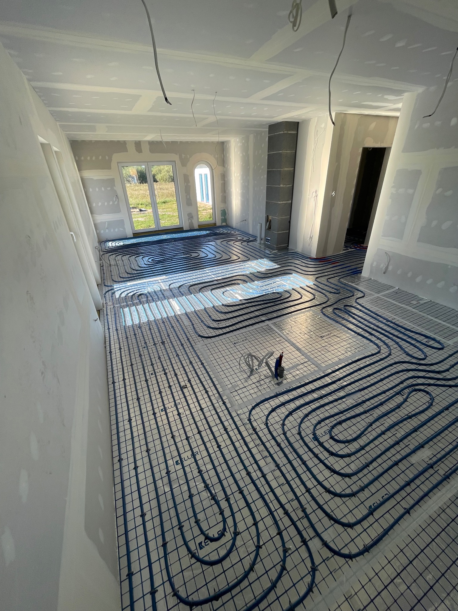 Instalacja ogrzewania podłogowego z niebieskimi rurami na szarej macie, widok z góry w surowym wnętrzu z oknami i przewodami.