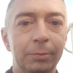 Portret mężczyzny z bliska, z krótkimi włosami i ciemnym ubraniem. Widoczne zmarszczki wokół oczu i delikatny uśmiech. Tło rozmyte, jasne.