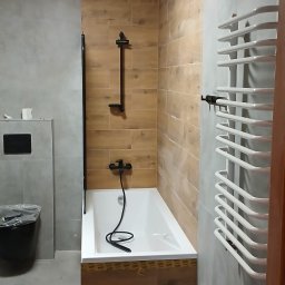 Angelika Krause - Wykończona łazienka z wanną obudowaną drewnopodobnymi płytkami, szara ściana, biały grzejnik drabinkowy i czarna armatura. Widok zza framugi drzwi.