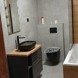 Angelika Krause - Wykończona łazienka w szarościach z czarną armaturą: umywalka nablatowa, toaleta podwieszana. Częściowo widoczna wanna z drewnianym wykończeniem.